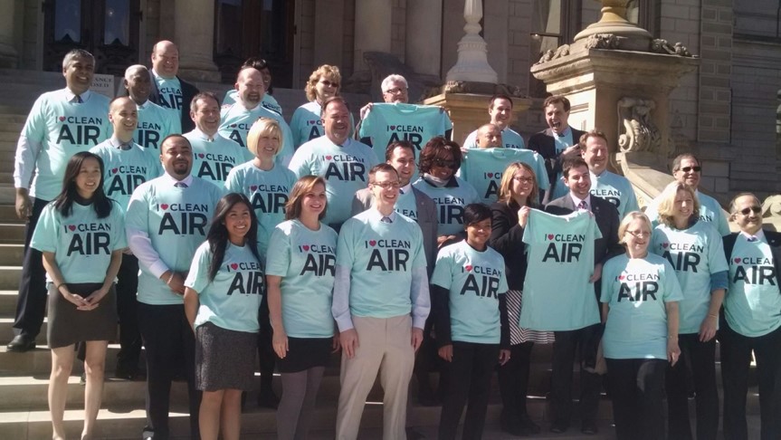 A crowd of about 30 people in matching blue, "I ❤ Clean Air" pose for a photo in front of a civic building. A breadth of ages, genders and races are present, and no one isn't smiling.