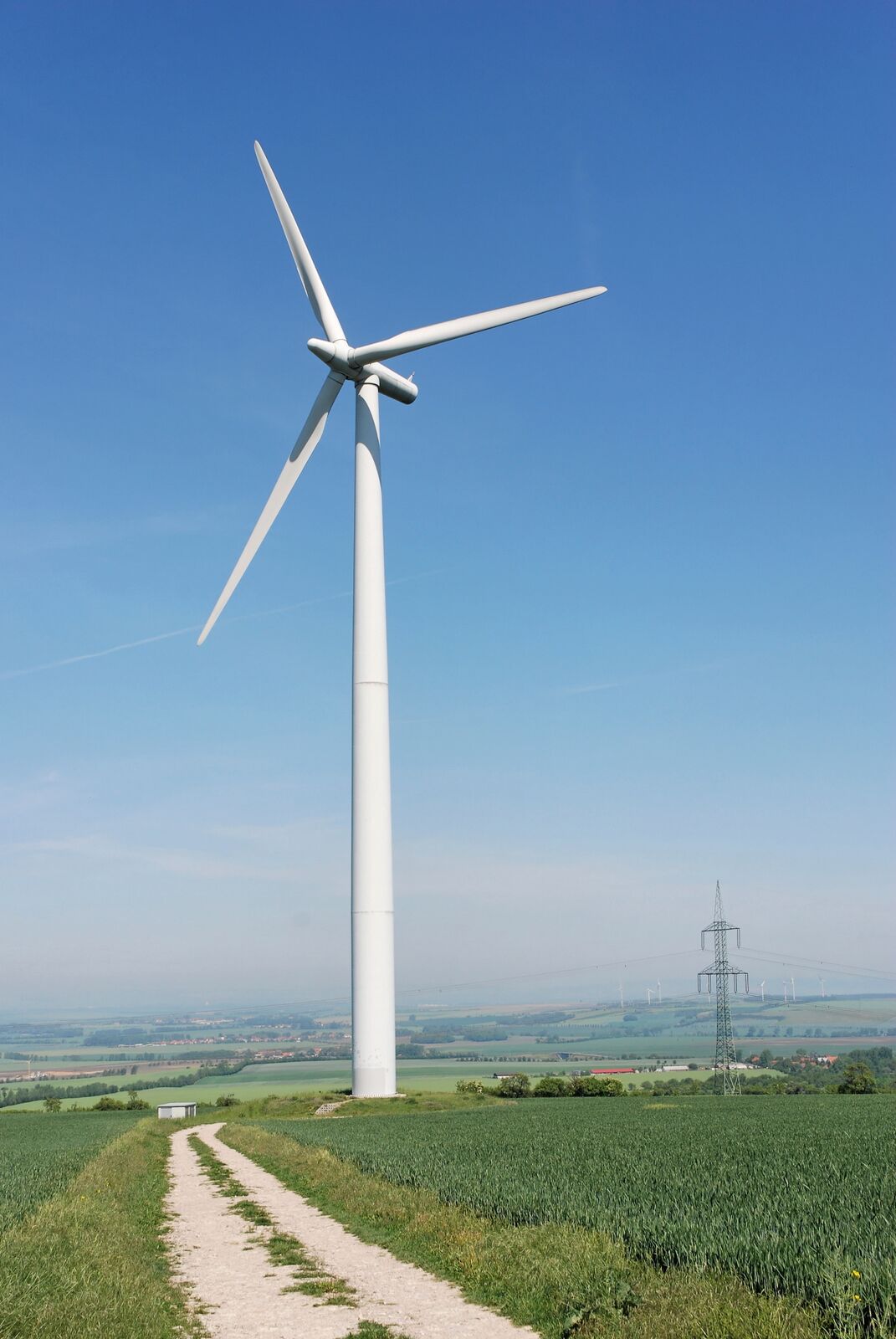 A small country path winds past a wind turbine towering above a rural panorama on a clear, sunny day
