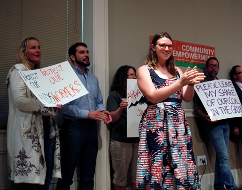 A smiling crowd stands mid-applause. In background, two signs can be made out: one reading "Protect our Kids, Protect our Workers" toward the left and toward the right, "PLEASE LEAVE MY SHARE OF OUR COAL IN THE GROUND."