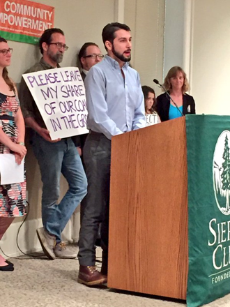 A bearded person stands before a podium displaying Sierra Club's banner depicting its pre-2015 logo. Behind the person stands a crowd of various ages and genders, one holding a sign that reads, "PLEASE LEAVE MY SHARE OF OUR COAL IN THE GROUND."