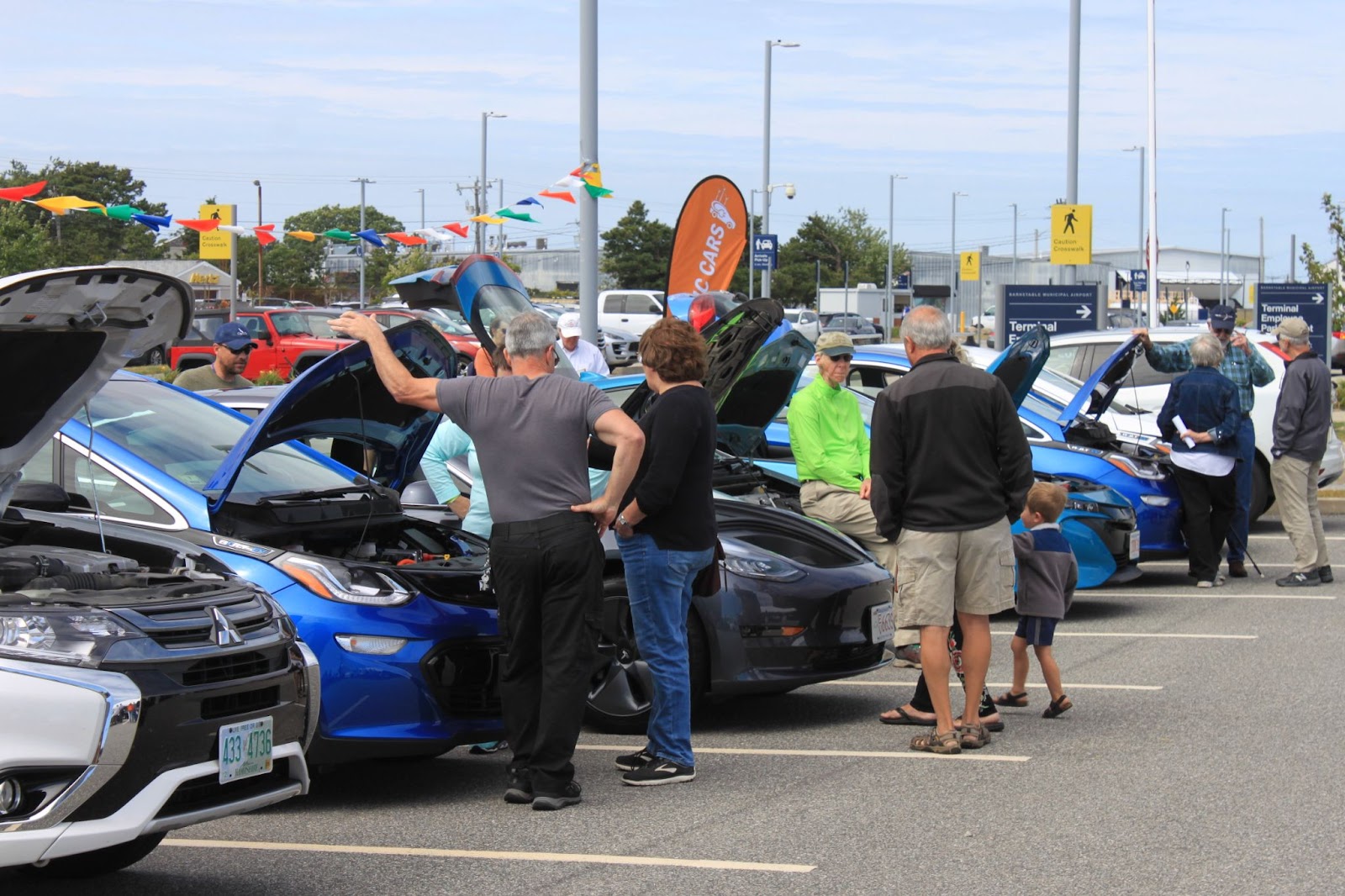 A line of electric vehicles with their hoods open, and various people looking at the vehicles and speaking with each other