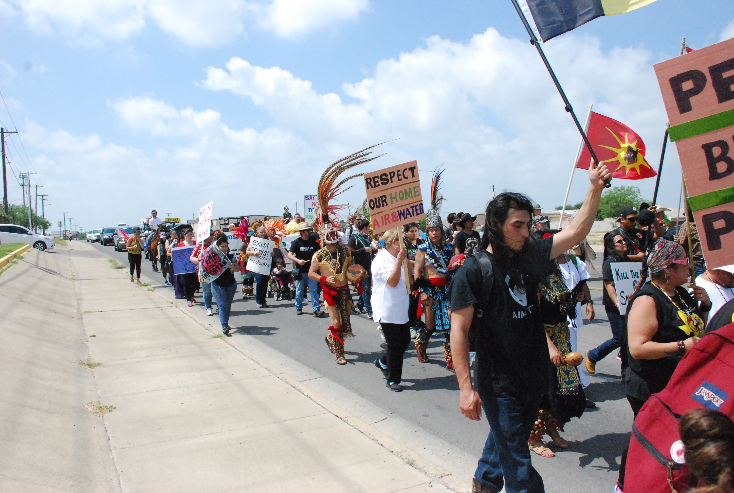 A portion of a large crowd of marchers holding signs like, "Respect Our Home, Air, and Water," "exist & resist... & decolonize," and, "Say No to the Mine." Crowd is predominantly indigenous, with dancers in regalia moving among the marchers