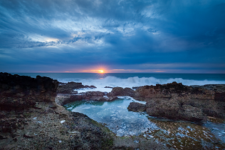 Sun at horizon of ocean with rocky coast and pool in foreground