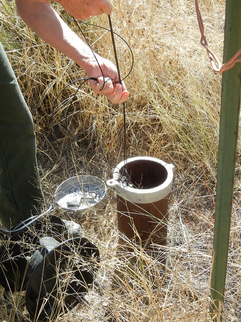 Two hands lowering a probe on wire into a small groundwater monitoring well in a dry, grassy area