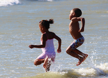 Small girl and boy in swimsuits leaping over waves at the beach on a sunny day