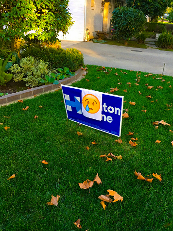 A Clinton-Kaine yard sign in a lawn that has been covered by a smaller sign with a crying face emoji on it.
