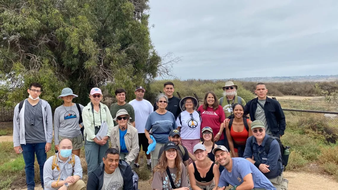 A group of 20 hikers poses at the Bolsa Chica Ecological Reserve, smiling for the camera, with trees and wetlands in the background. 