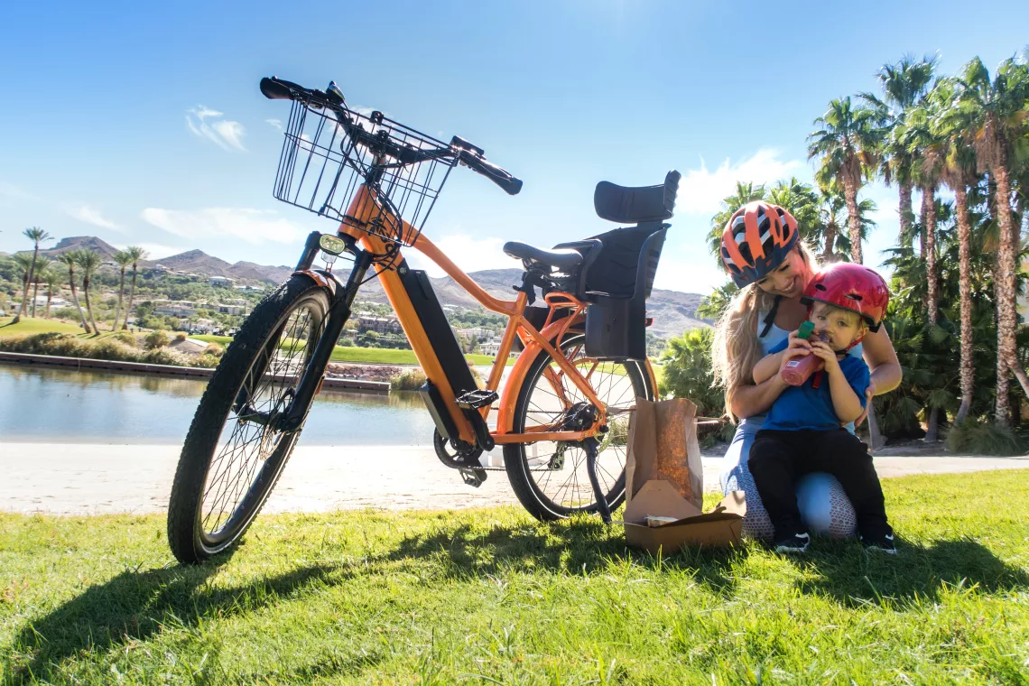 Mother and son wearing helmets and sitting next to an e-bike on some sunny grass.