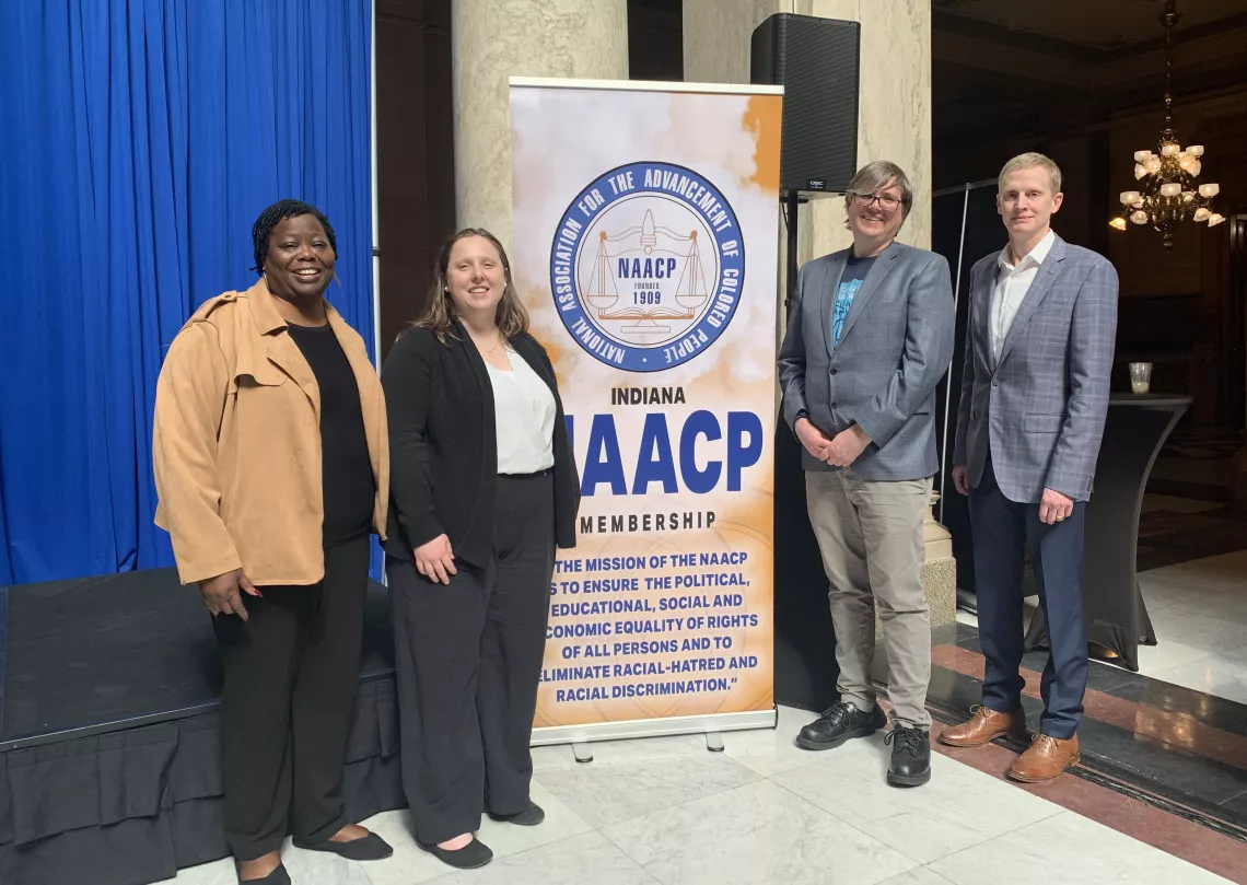 Four people stand at an indoor venue, smiling for the camera. They are standing by an NAACP banner.