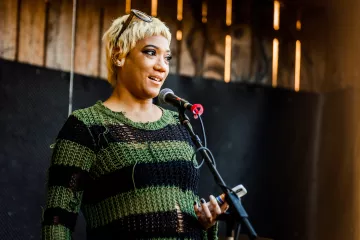 A woman with short blond hair and a green and black stripy sweater stands at a microphone reading poetry.