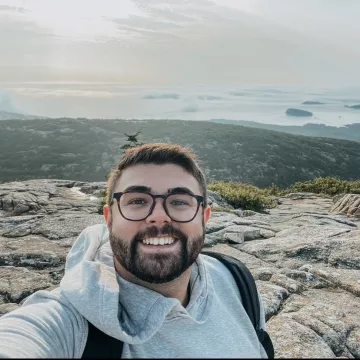 A person with short brown hair, glasses, and a brown beard smiling for a selfie. He is wearing a gray hoodie and a backpack. He is standing on top of a mountain.