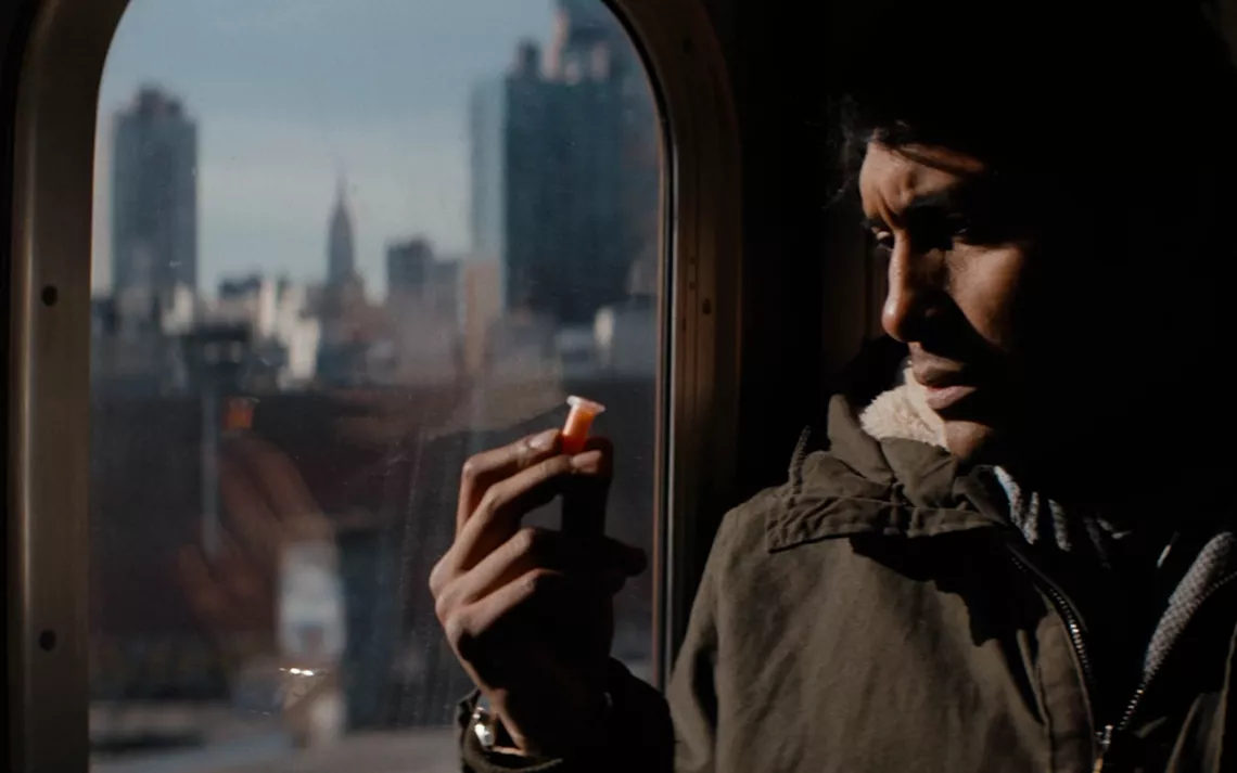 A man sits on an MTA train as it crosses over a watery landscape, looknig at an orange vial.