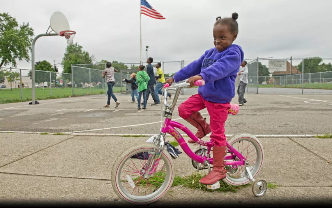 Myshelle James, 4, rides her bike in River Rouge's Memorial Park while her brothers play basketball in the background and her mother, Alisha Winters (not shown), helps at a nearby farmers' market. | Ami Vitale/Panos Pictures