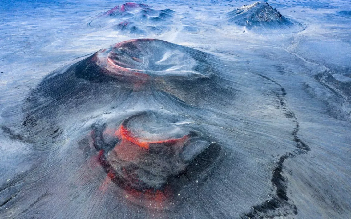 The snow dusted cone of a volcano, with red hot and gold magma contrasting with the blue ice and snow 
