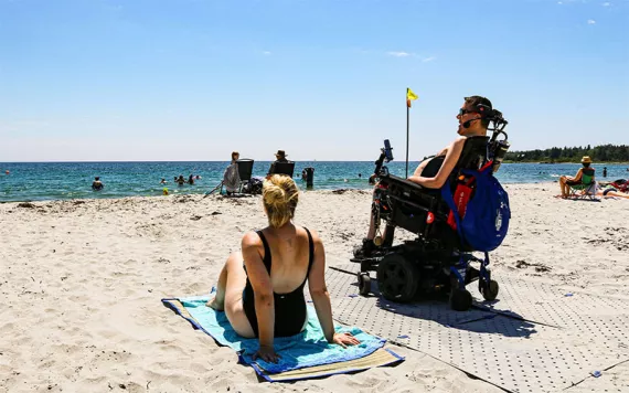 A person sits in a wheelchair on the beach, next to another person sitting on a towel on the sand.
