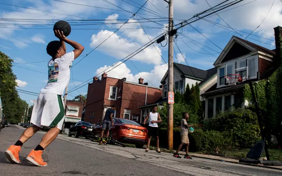 A young man prepares to take a shot at a basketball hoop set up on the street in front of houses in Pittsburgh. Four other young men stand around.