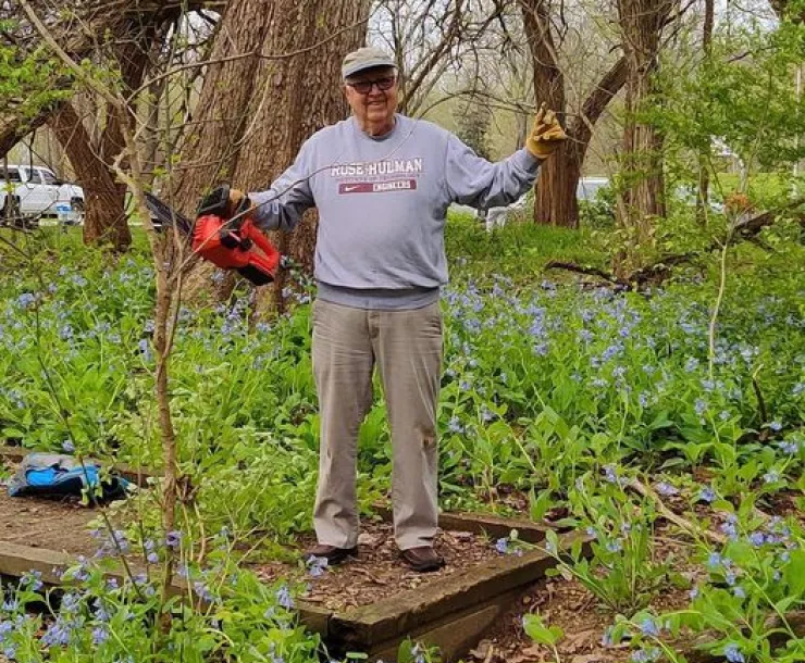 A senior gentleman stands with a chainsaw in his hand surrounded by invasive honeysuckle which is a short green plant. He is smiling.