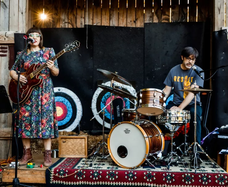 Two people perform on a small wooden stage outdoors. One is a woman wearing a floral dress playing guitar and singing. She is standing. The other person is a man seated and playing drums and singing.
