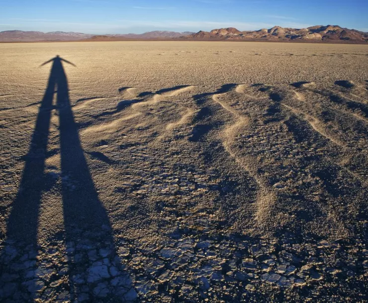 A person's tall black shadow stretches far across a desert plain into the distance. Waves in the sand point toward brown mountains in the background.