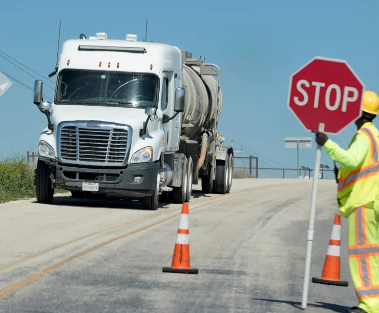 A white 18 wheeler truck moves down a road with an employer in safety gear holding up a stop sign