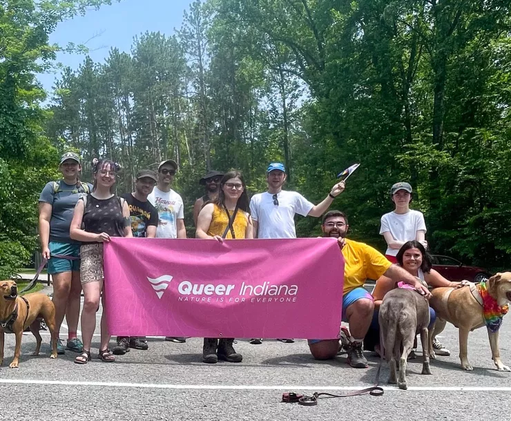 A group of people standing in a parking lot with trees behind them on a sunny day. There are a few dogs present too. A few of the people are holding up a pink banner saying Queer Indiana in white writing. They are smiling.
