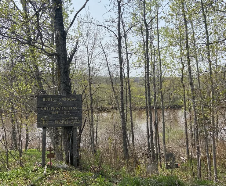 Image of a sign identifying an Ojibwe burial ground in St. Francis Cemetery, with the Nemadji River in the background.