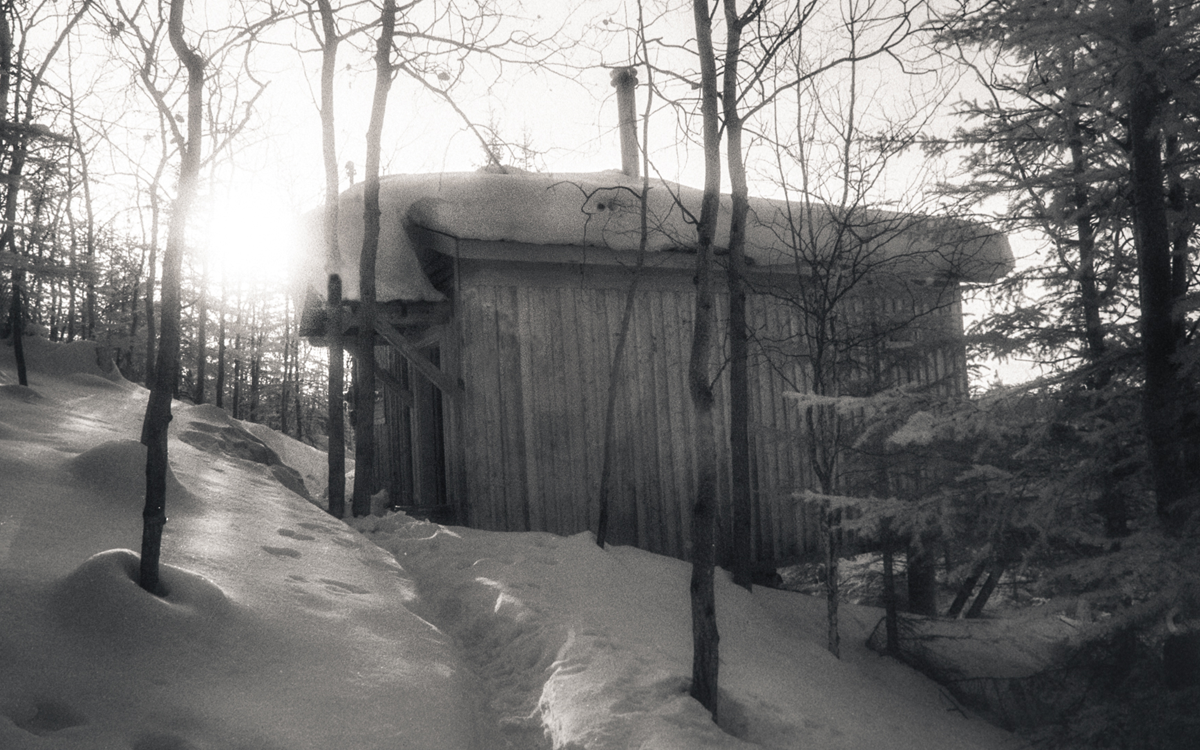 A black-and-white photo shows John Luther Adams's Alaskan cabin perched on a hill in the snow.