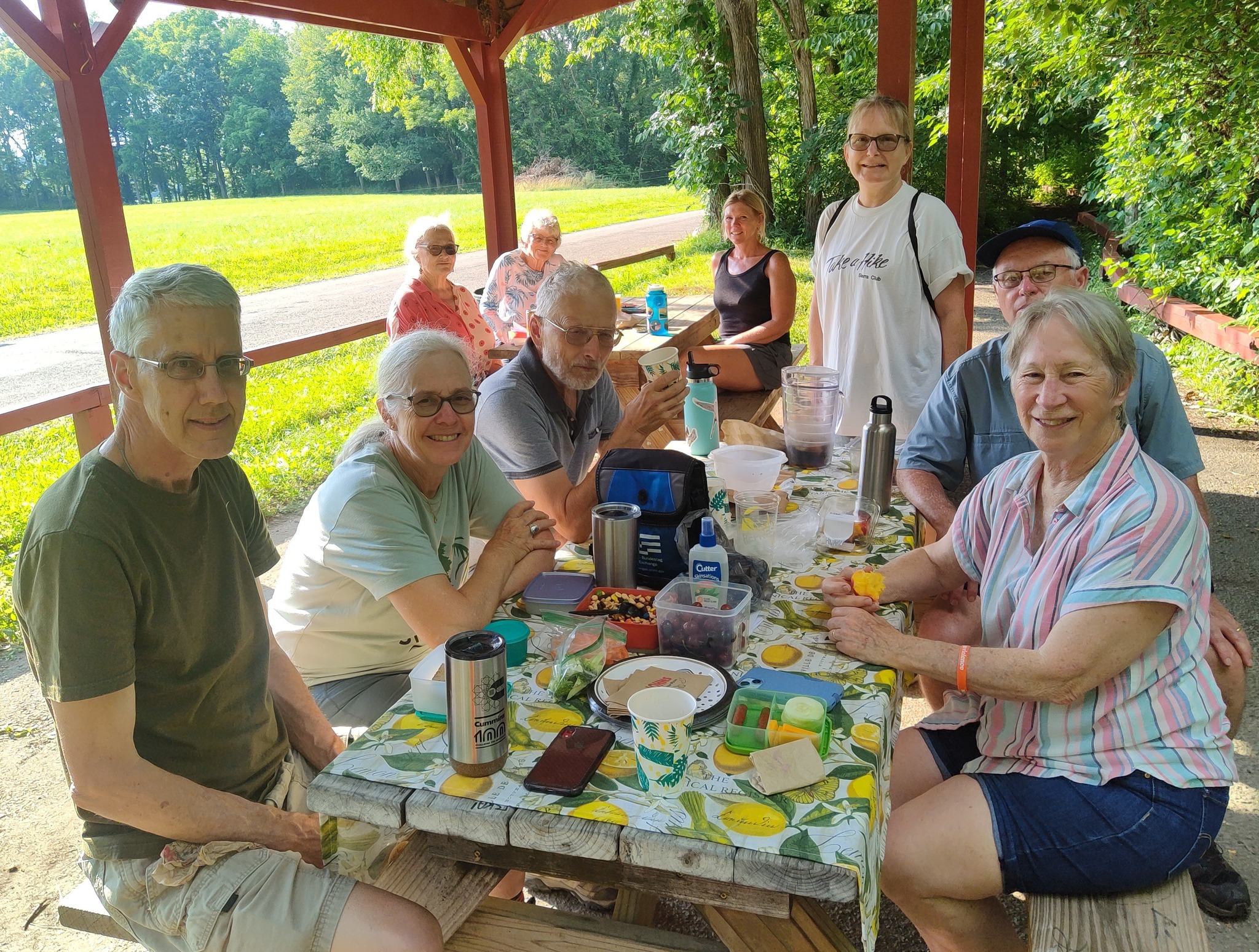 A group of nine people sitting outdoors eating at an outdoor picnic table. They are looking at the camera and smiling.