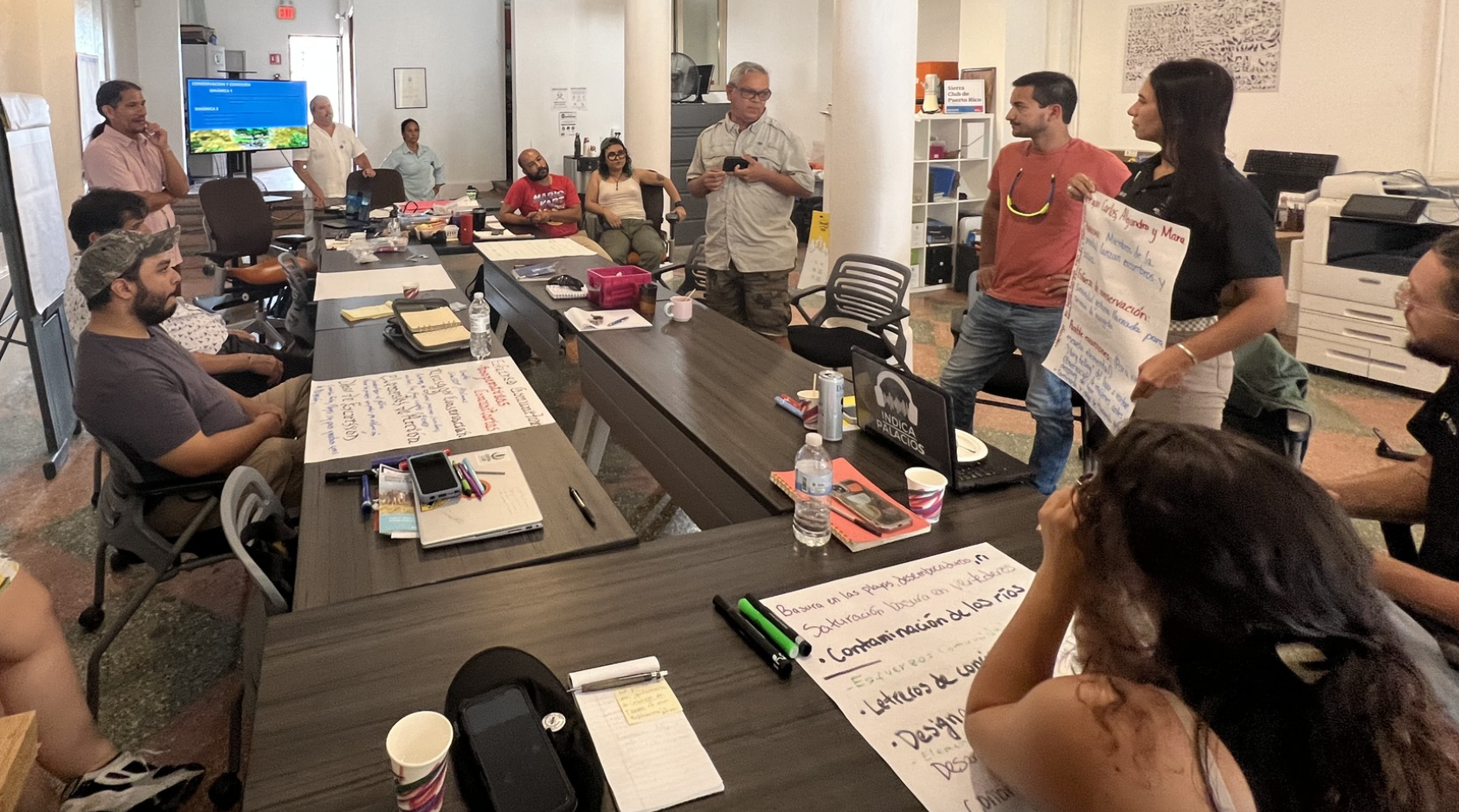 A group of people sit and stand around a table, collaborating and sharing ideas, with papers and materials spread out as one person presents to the group.