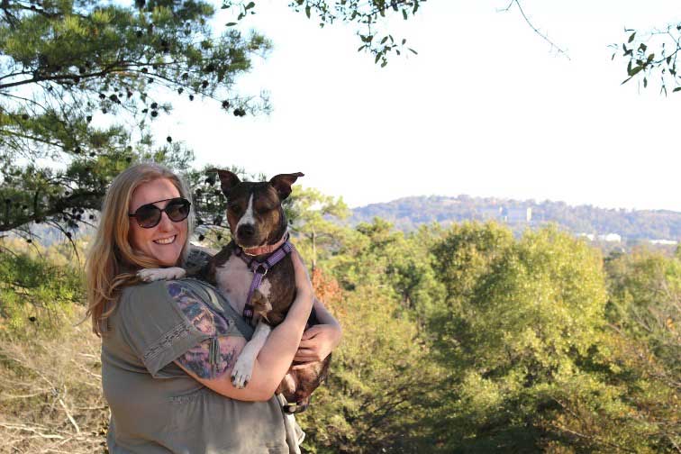 Emily Bosch, white woman wearing sunglasses and holding her dog with a pine tree and hills in the background
