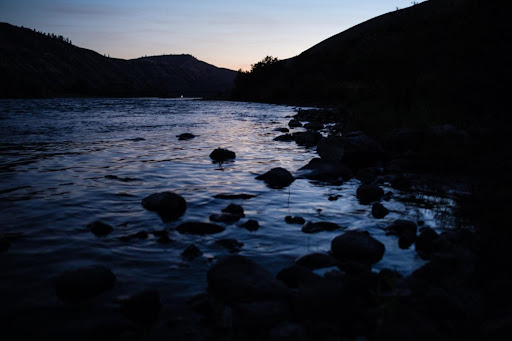The Snake River at sunset is a mix of pale blues and pinks, reflecting the sky. The surrounding hills, shore, and rocky outcrops are silhouetted in black
