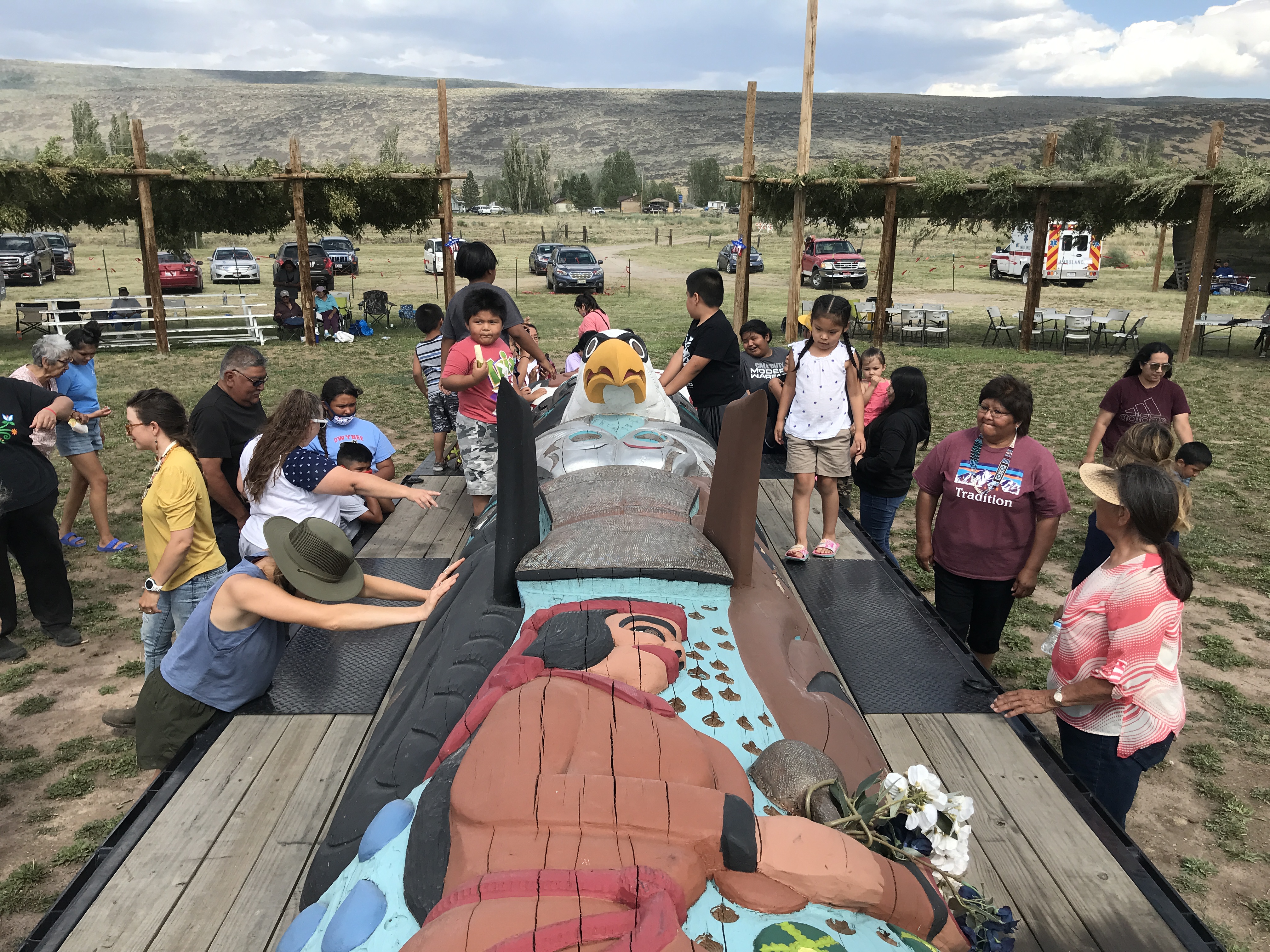 Native people, including children, gathered around an intricate totem pole lying on its side on a truck bed, at the Duck Valley Shoshone-Paiute Reservation in Nevada