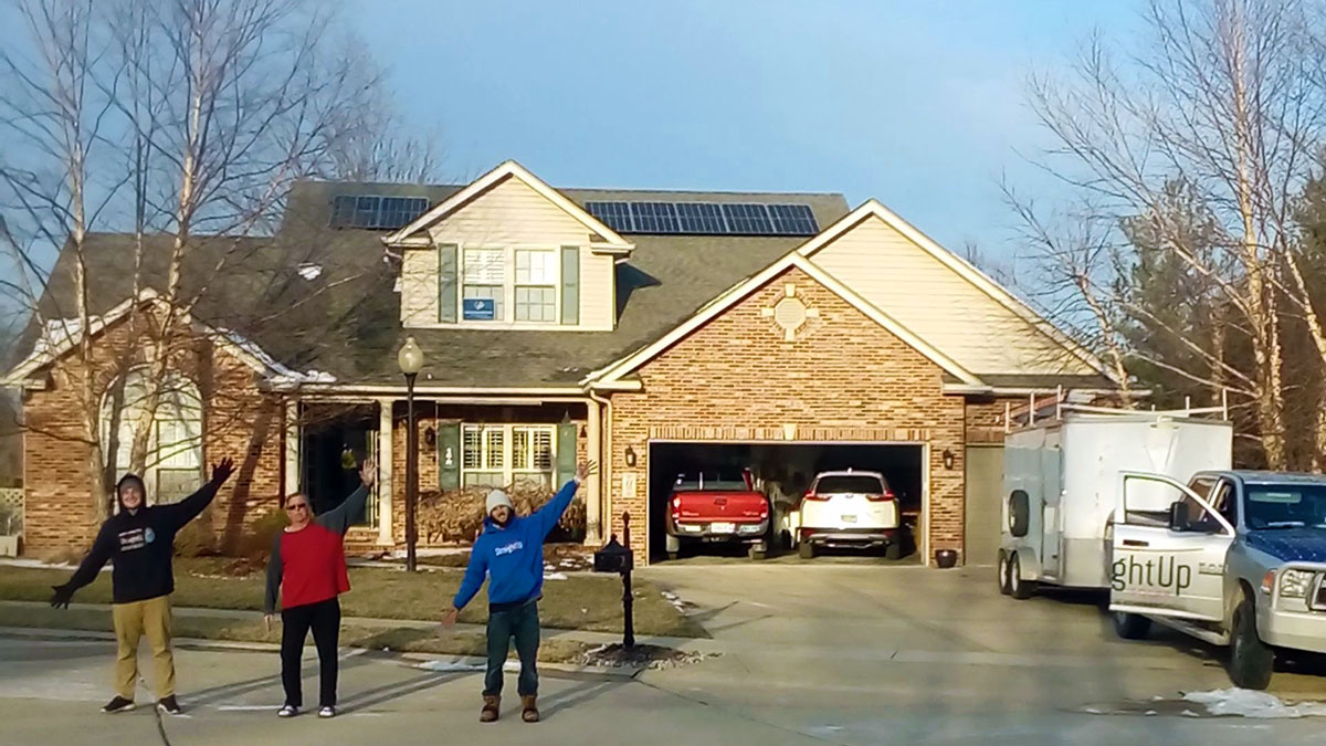 Three people stand in front of their home, which features solar panels on the roof.