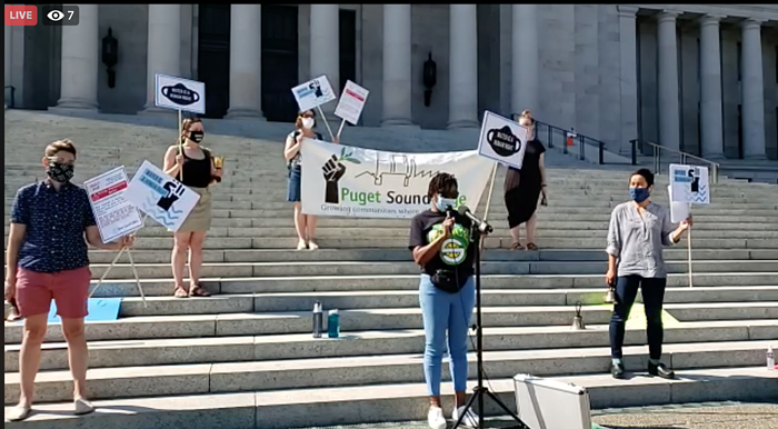 A group of organizers standing on the steps of the Olympia Capitol. They are holding signs as Yolanda Matthews from Puget Sound Sage asks Gov. Jay Inslee to "lessen the burden" on low-income families.