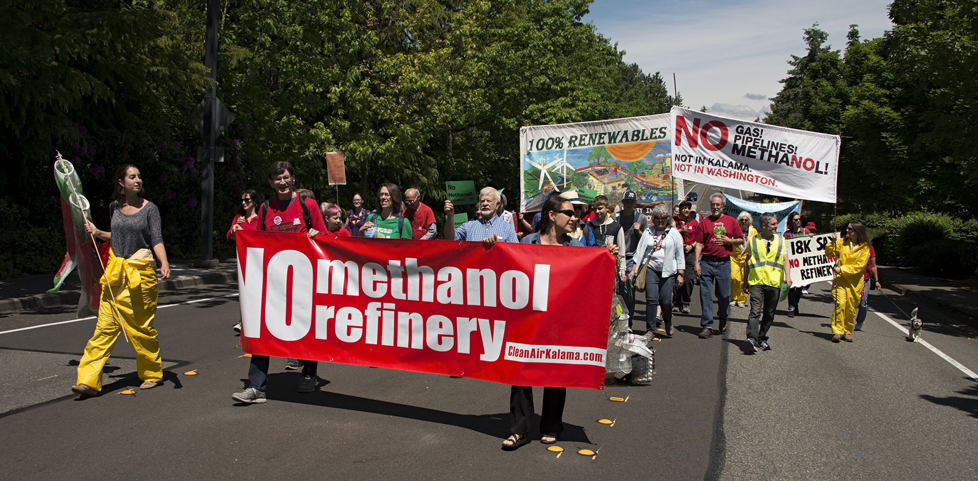 A group of protestors marching down the street holding a large banner between them. The banner is red with white letters and it reads: No methanol refinery! It has a link on the bottom right corner for CleanAirKalama.com.