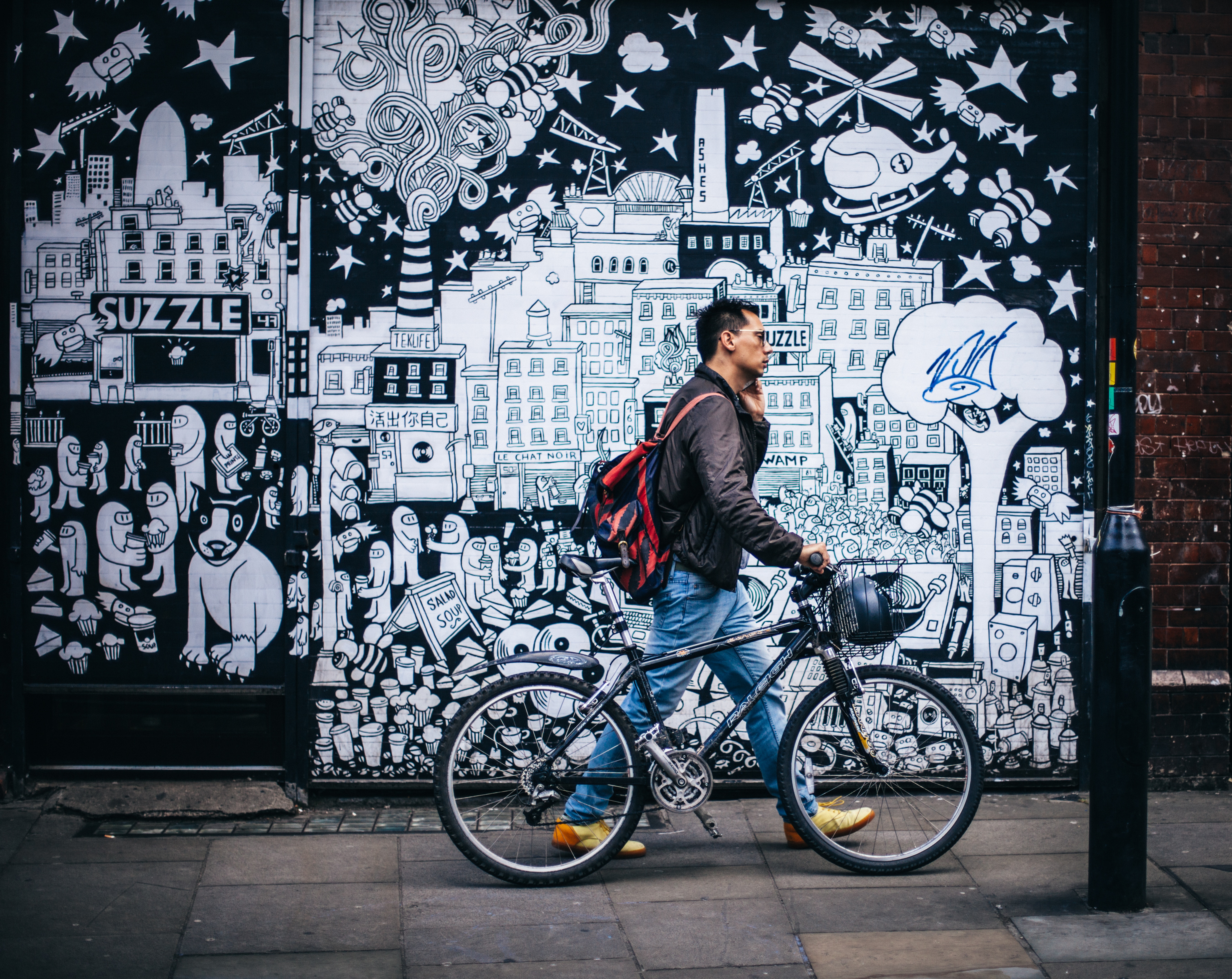 Man with red backpack talking on phone while guiding bike on the sidewalk. In the background, a wall of neat graffiti. 