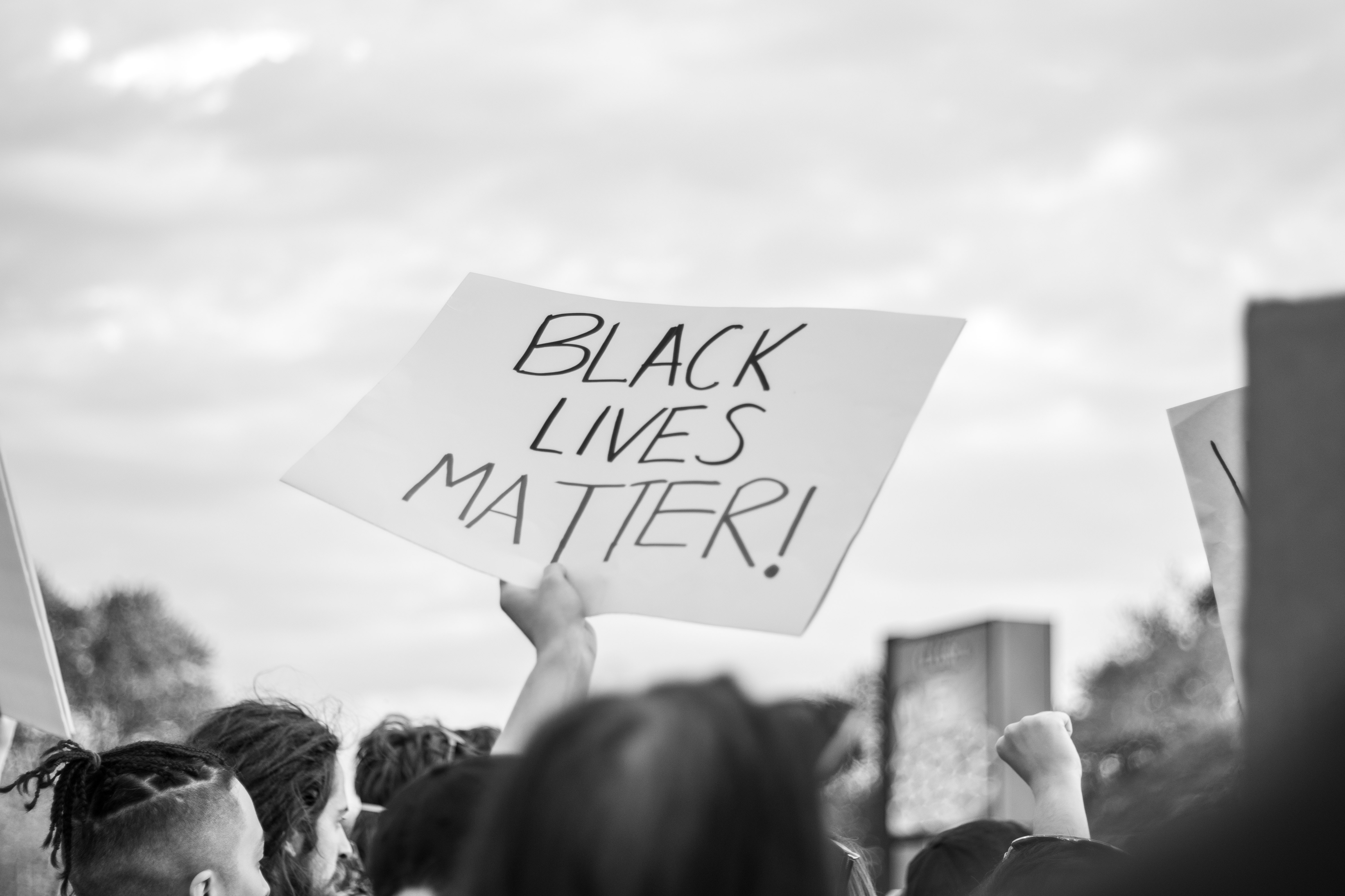 A hand holding up a sign amidst a crowd. The sign reads " Black Lives Matter!"