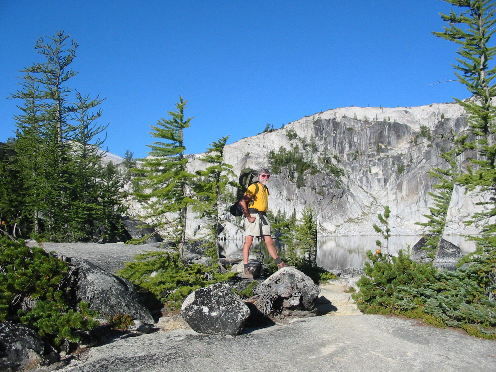 dick viviane stands in a yellow short and beige shorts against a bright blue sky and green trees and white boulders.