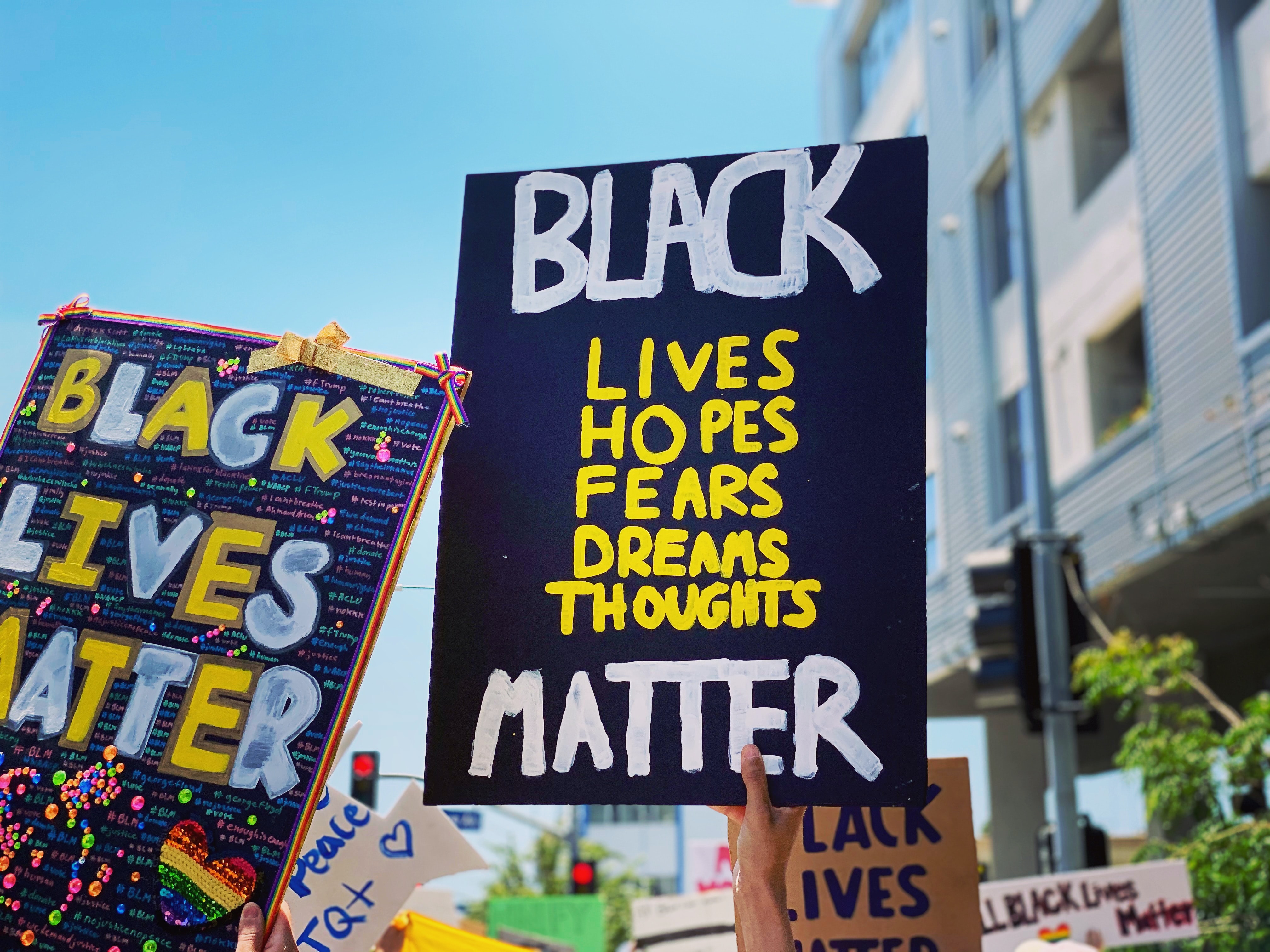 A black sign being held aloft. It reads: Black Lives, Hopes, Fears, Dreams, Thoughts Matter." Next to this sign is another one that reads "Black Lives Matter"