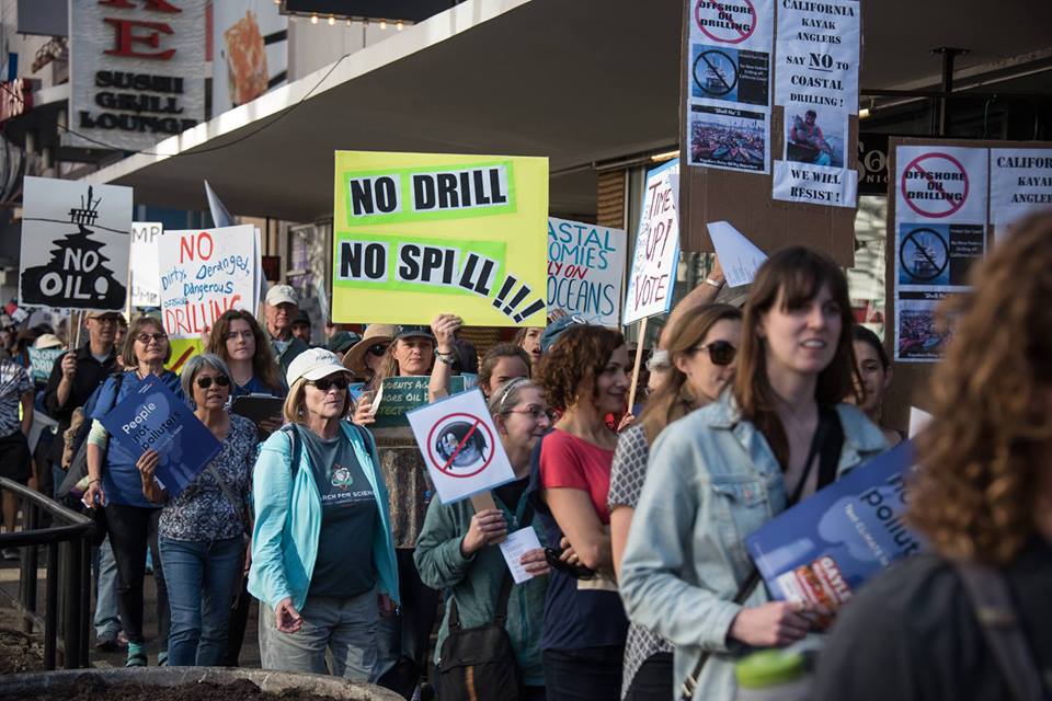 A group of people walk along a sidewalk holding signs, one of which reads No Drill, No Spill