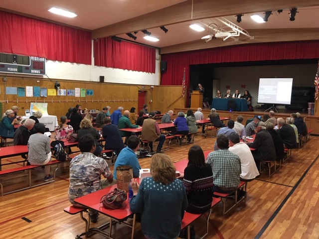 People sit around cafeteria tables in a school auditorium while watching a presentation