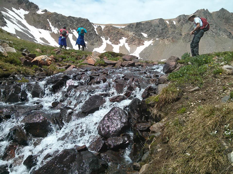 A waterfall tumbles over rocks in the foreground, as three hikres stand at the top looking at mountains in the background.