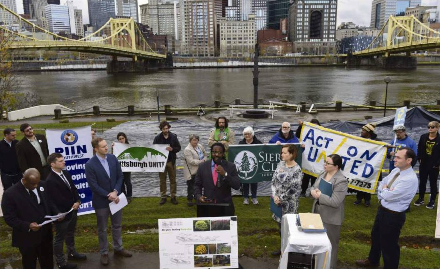 A crowd gathers around a speaker in support of clean rivers, with two yellow bridges over the Allegheny River in the background.