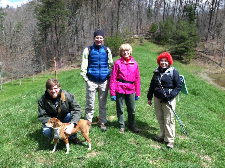 There are women and one man standing in hiking gear on a grassy hilltop. A second man is squatting and petting a dog.  