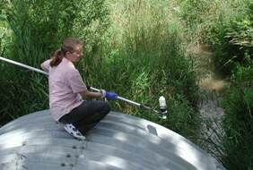 A volunteer reaches down from atop a culvert to take a water sample from a stream.