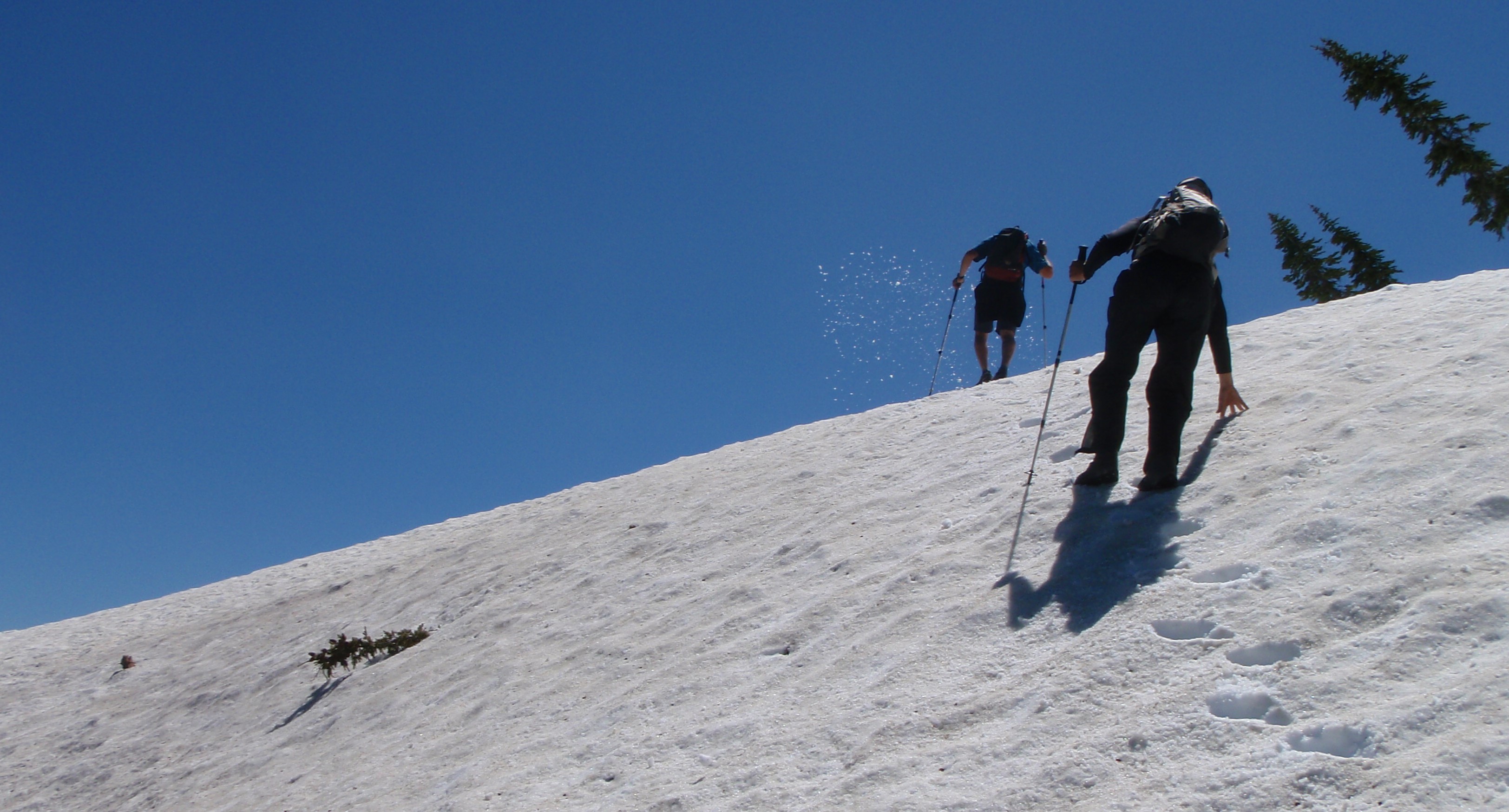 Two people hike across a snowfield with the sky behind them.