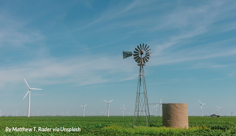 A windmill and water tank stands in the foreground of a picture of a wind farm in West Texas on a sunny day