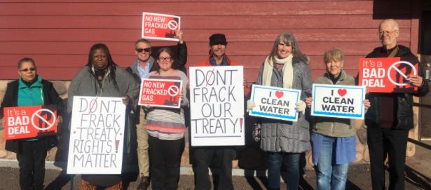 Photo showing eight people standing in front of a red building holding signs that oppose new gas