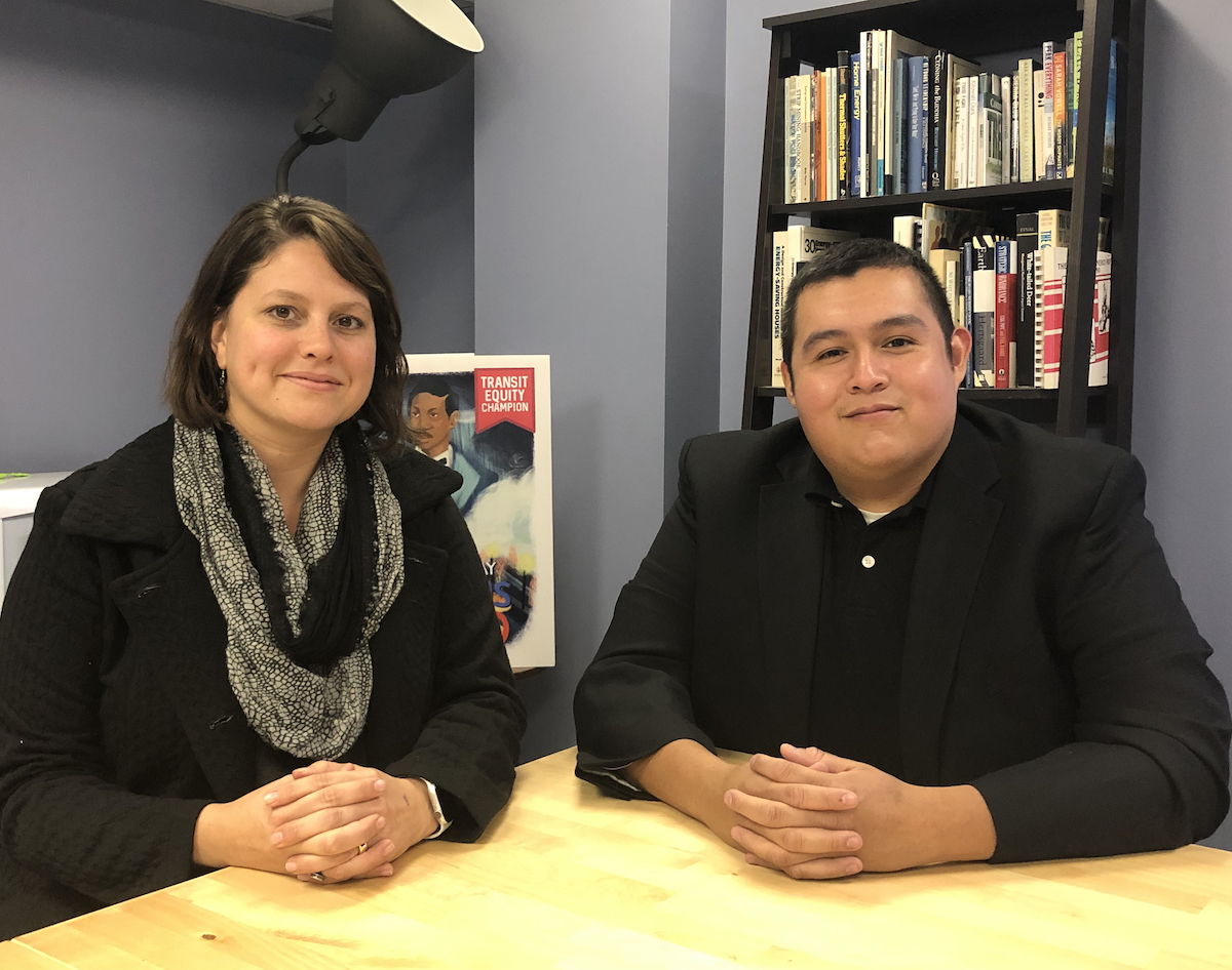 Two people, Joanne and Tom, smiling, wearing black clothes seated together at a conference room table. Behind them is a poster that says “Transit Equity Coalition”, along with a bookshelf