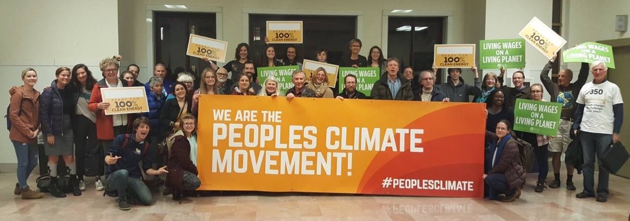 Dozens of volunteers of all ages gather with an orange "We are the People's Climate Movement" banner, "Living Wages on a Living Planet" and Ready for 100 signs after testifying in front of council for a just Climate Action Plan with an 100% renewable energy goal